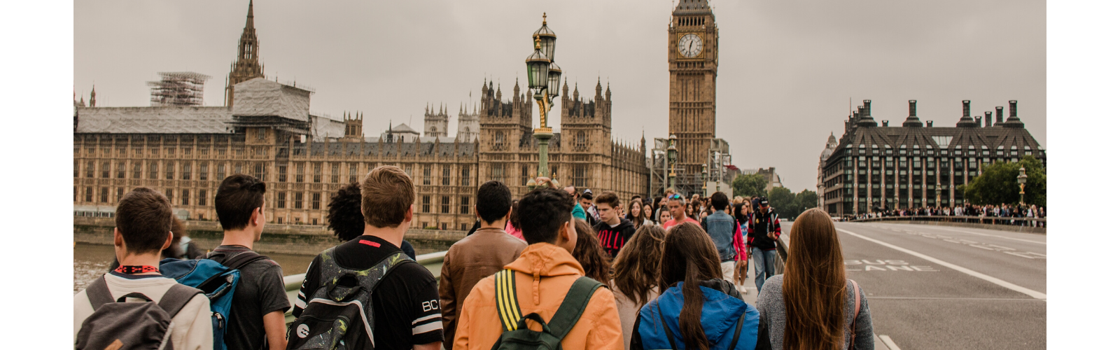 A group of students with backpacks are walking down a street with many pedestrians walking towards them. An extravagant palace and tall clocktower is visible in the background. 